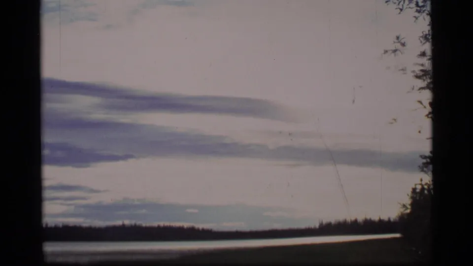 1977 Alaska 8mm frame of a quiet lake scene, with forest shoreline, overcast sky, and roadside foliage visible at frame edge.