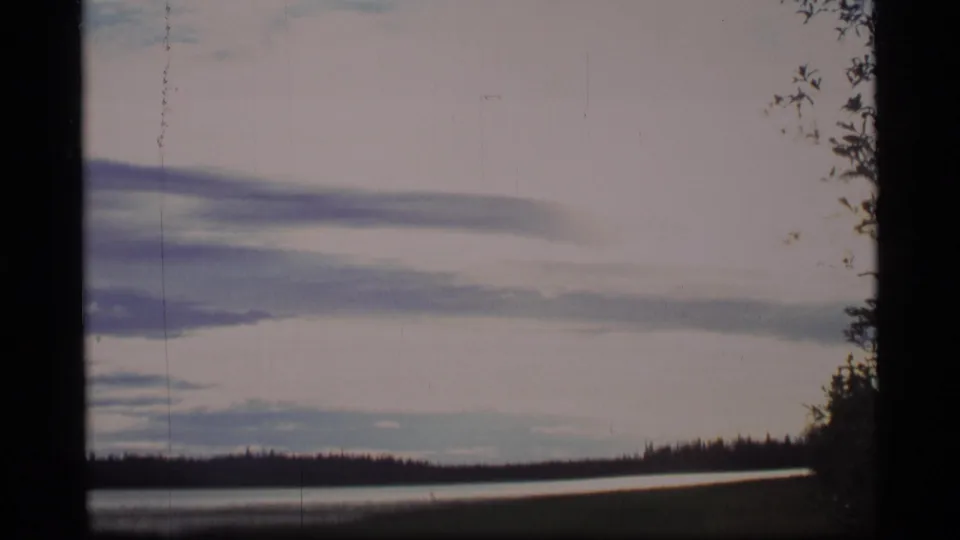 1977 Alaska 8mm view of lake water and a dark evergreen shoreline beneath a soft, clouded northern sky.