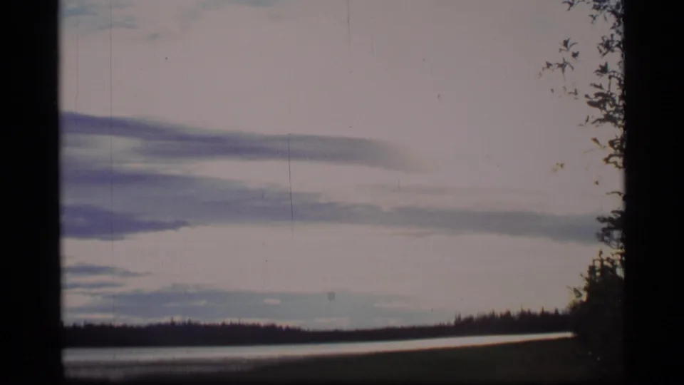 1977 Alaska 8mm frame showing an overcast lake view, low wooded horizon, and trees along the right edge.