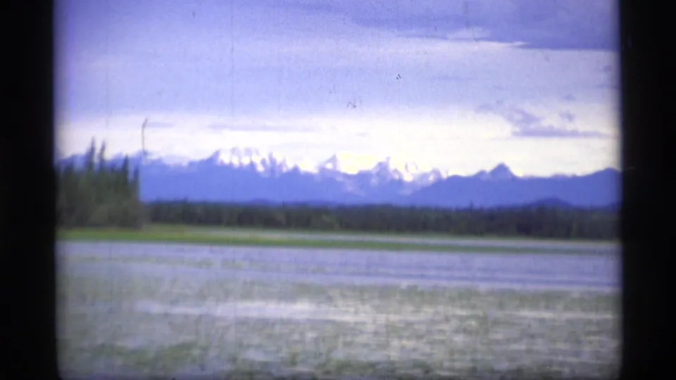 1977 Alaska 8mm frame looking across lake water toward forest and snow-capped mountains in the distance.