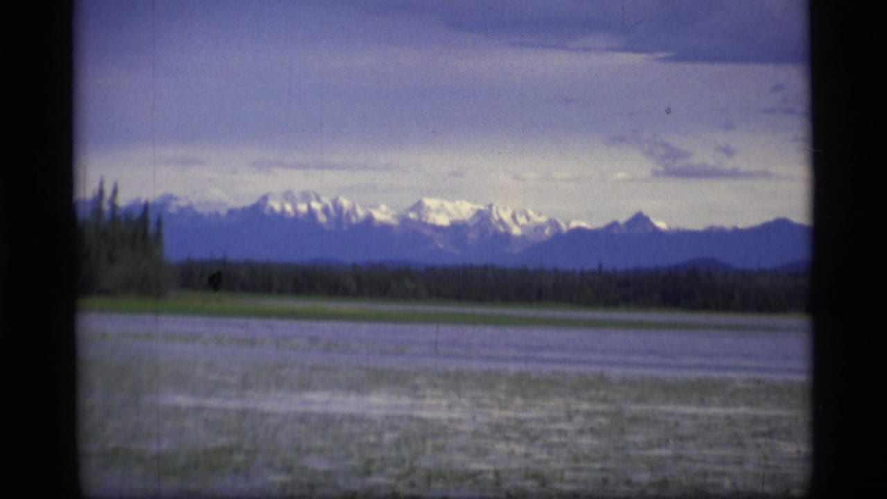 1977 Alaska 8mm frame showing a lake, dark tree line, and distant snow-capped mountains beneath a soft blue-gray sky.