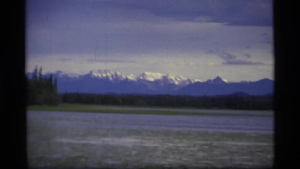 1977 Alaska landscape frame with a lake foreground, dark forest band, and snow-capped mountain range in the distance.