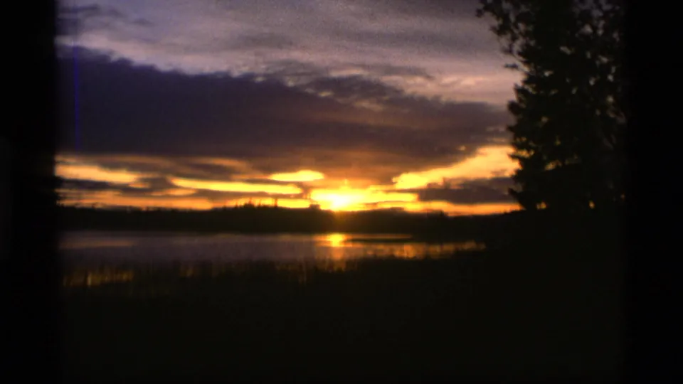 1977 Alaska 8mm sunset frame with orange light breaking through clouds and reflecting on water near silhouetted reeds and trees.