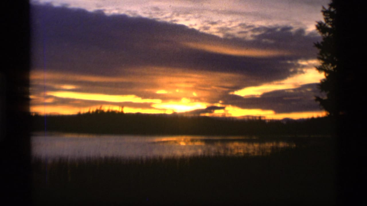 1977 Alaska 8mm frame showing a golden sunset reflected on lake water beneath layered clouds and dark tree silhouettes.