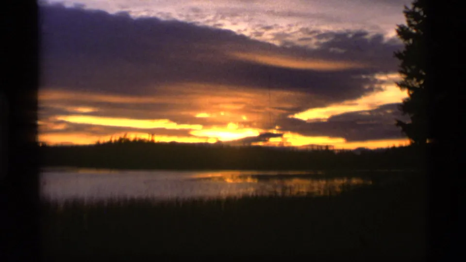 1977 Alaska home-movie frame with orange sunset light, cloud bands, reflective water, and silhouetted lakeside trees.