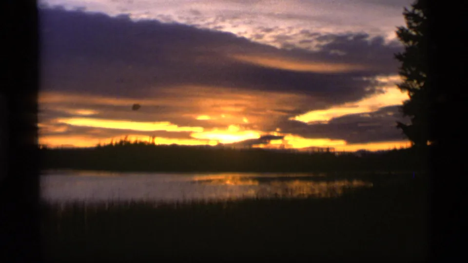 1977 Alaska 8mm frame of a low golden sunset behind clouds, with lake reflections and dark reeds in the foreground.
