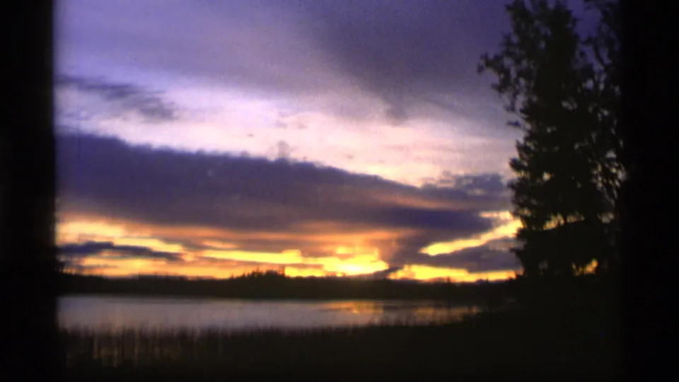 1977 Alaska 8mm frame showing a silhouetted evergreen beside lake water and a bright sunset band under violet clouds.