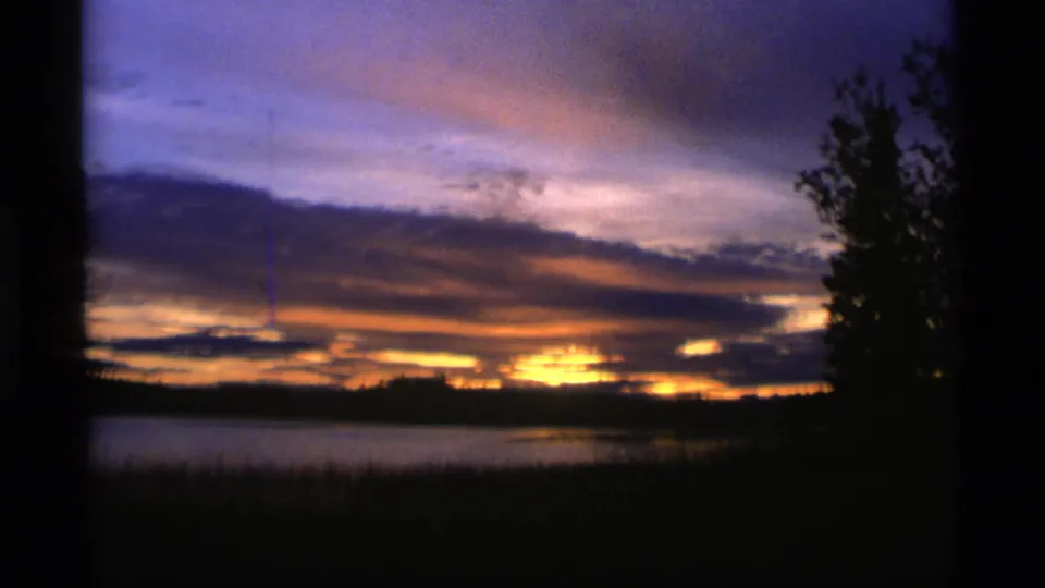 1977 Alaska home-movie frame with a broad sunset sky over a lake, dark shoreline, orange cloud bands, and a tree silhouette at right.