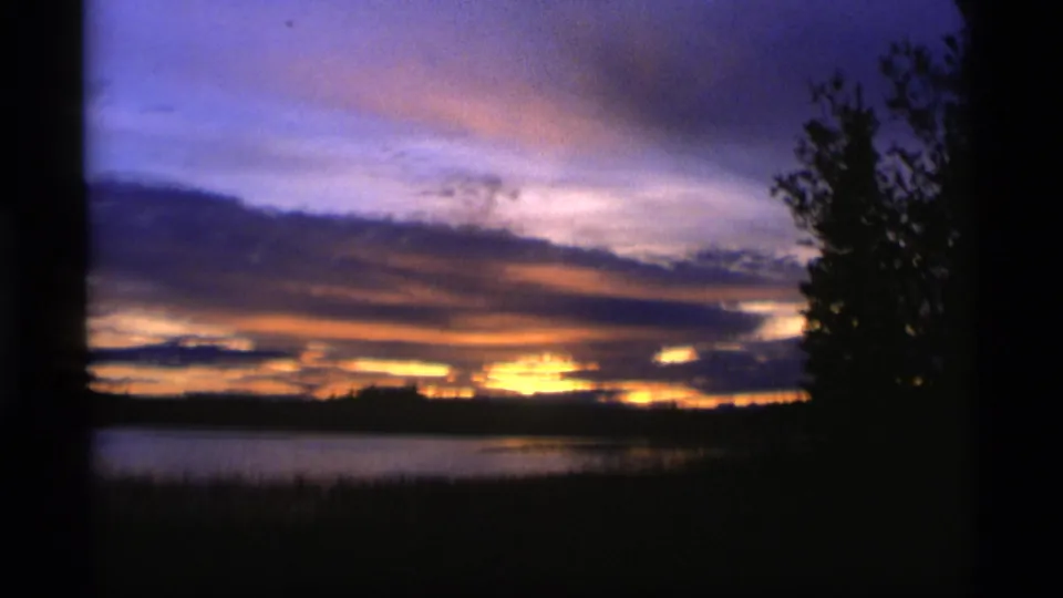 1977 Alaska 8mm frame showing a low gold sunset across a lake, with purple-blue clouds, silhouetted shoreline, and reeds in deep shadow.