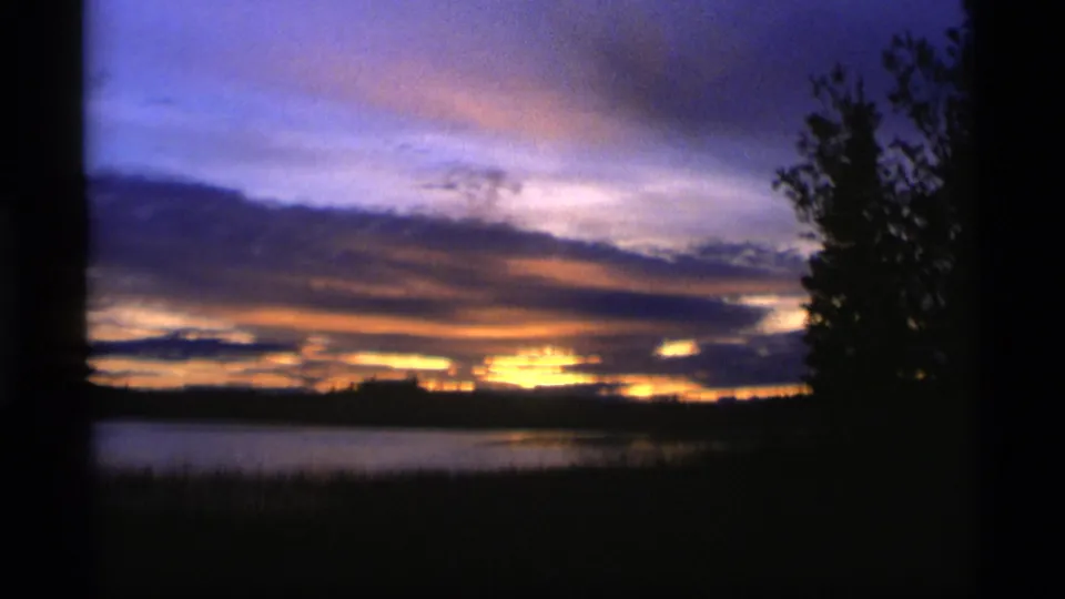 1977 Alaska 8mm frame holding on a twilight lake scene, with a gold band at the horizon, purple sky, and a dark tree mass at right.