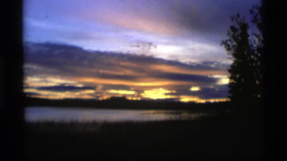 1977 Alaska 8mm frame with warm sunset light near the horizon, calm water, and silhouetted lakeside trees.
