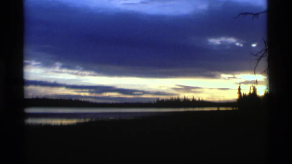 1977 Alaska 8mm frame showing a cooler twilight sky over water, with silhouetted shore trees and bare branches.