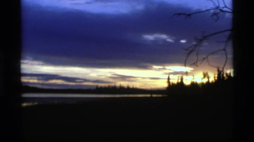 1977 Alaska 8mm frame of a lakeside dusk view with low clouds, bright horizon light, and silhouetted branches.