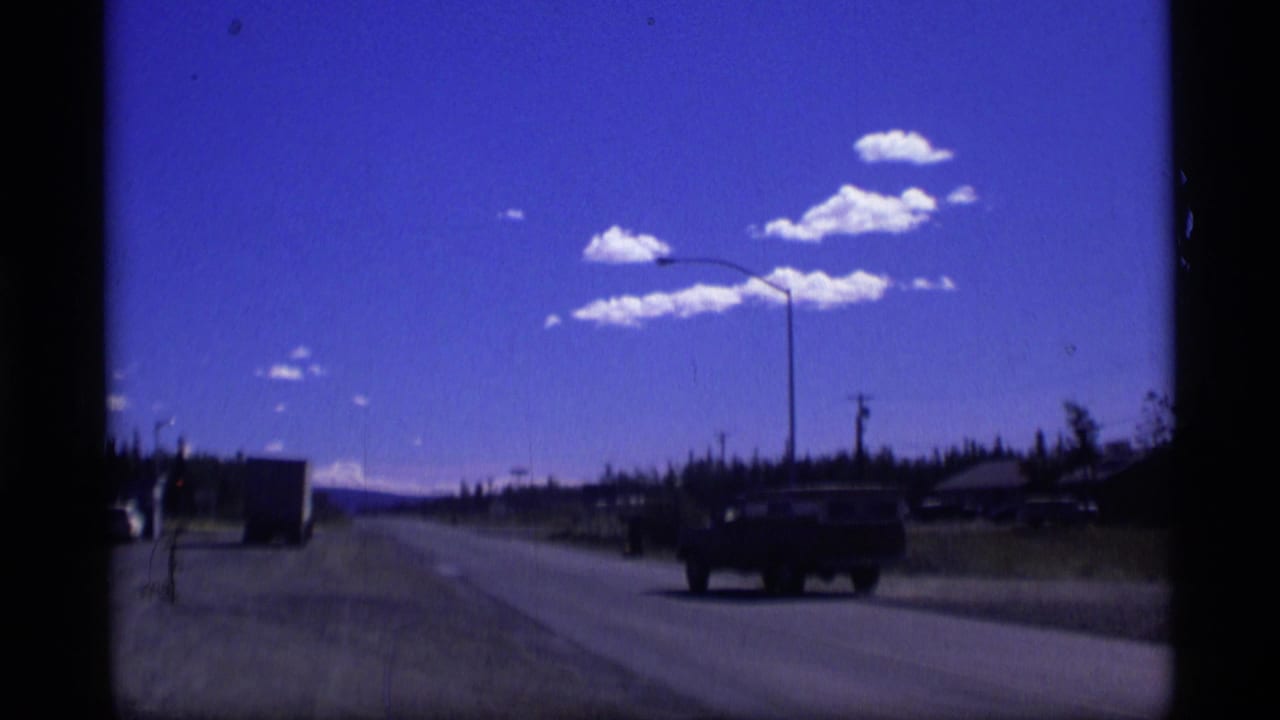 1977 Alaska 8mm road-trip frame showing a pickup truck on a dusty rural highway near roadside structures.