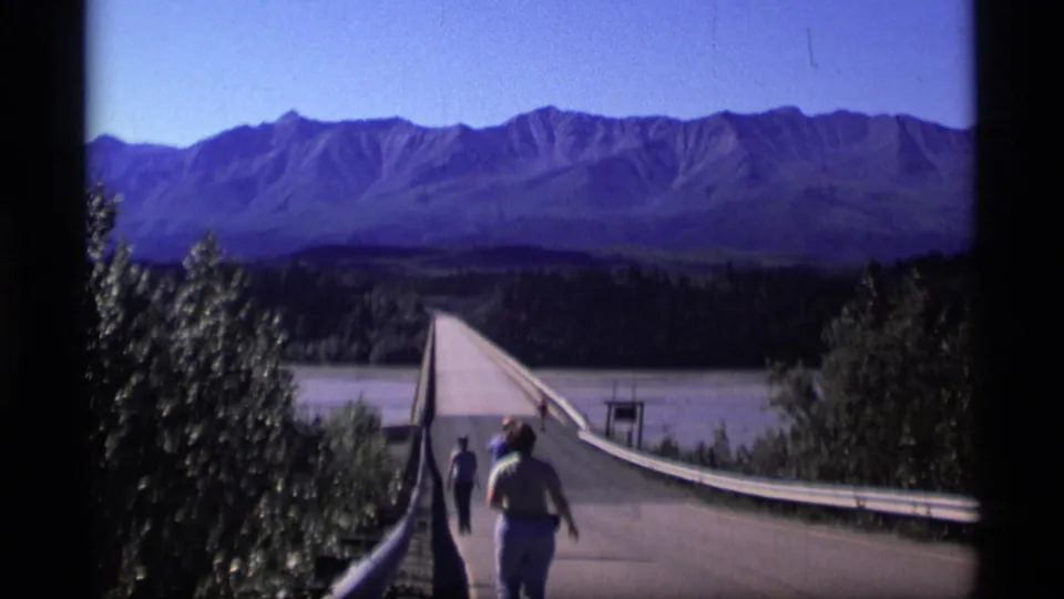 1977 Alaska home-movie frame of pedestrians on a long bridge crossing a river toward a mountain range.