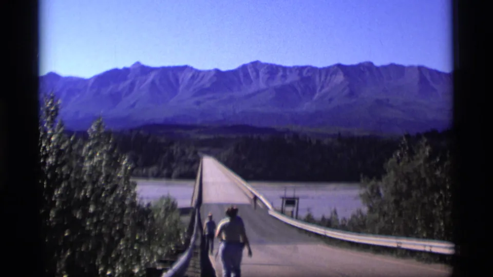 1977 Alaska 8mm frame showing two pedestrians on a bridge approach above a river with mountains beyond.