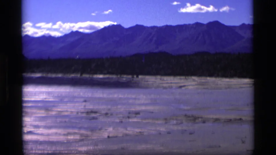 1977 Alaska travel frame showing a broad river or tidal flat below a dark mountain range and bright clouds.