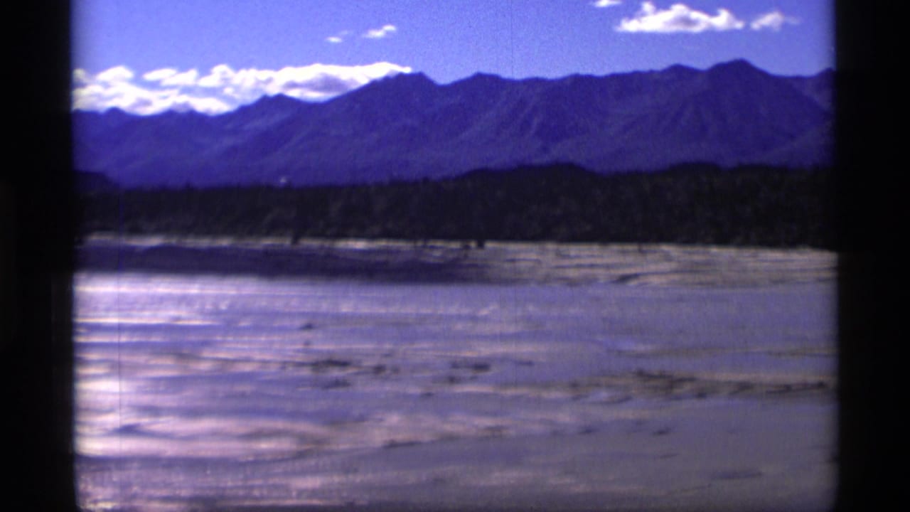 1977 Alaska 8mm frame showing a bright riverbed or sandbar area below a dark tree line and distant mountains under scattered clouds.