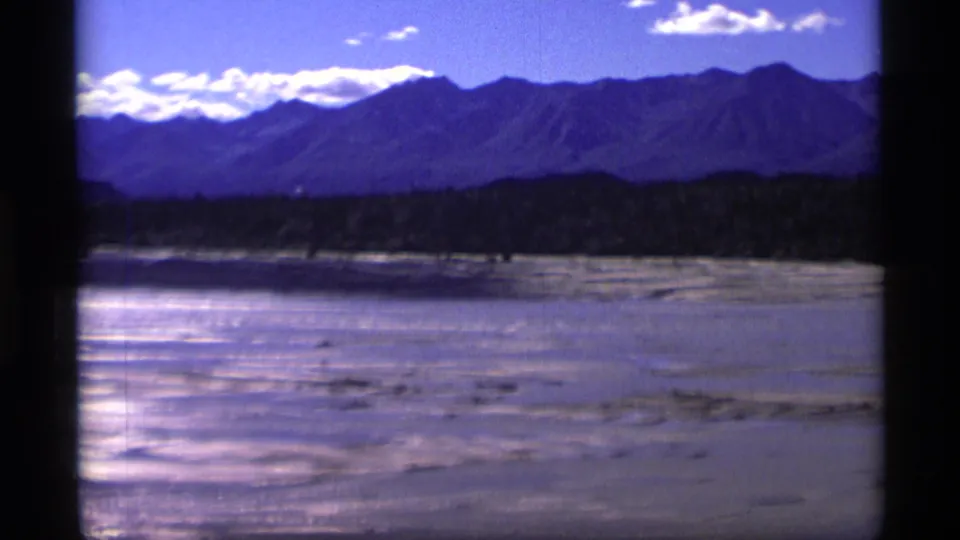 1977 Alaska 8mm frame showing a bright riverbed or sandbar area below a dark tree line and distant mountains under scattered clouds.