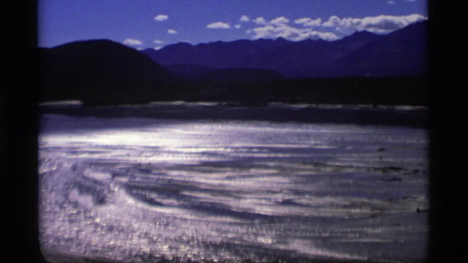 1977 Alaska 8mm frame showing braided-looking water channels and pale sandbars beneath dark hills and distant mountains.