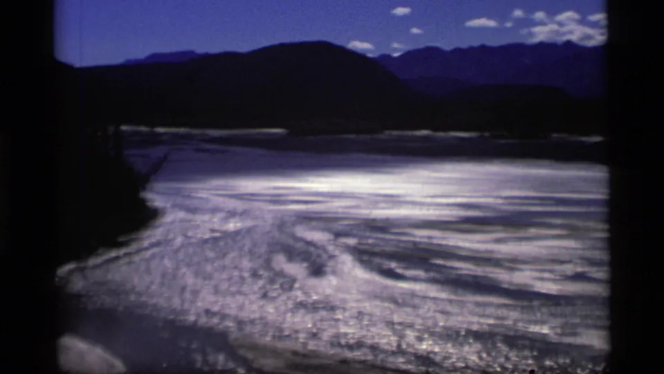 1977 Alaska 8mm frame capturing a curving reflective riverbed with exposed sandbars, dark hills, and mountains under a blue sky.
