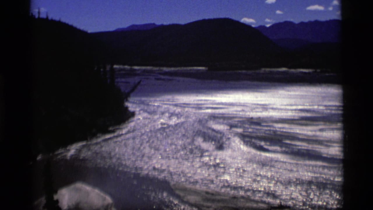 Blue waterway with sun glare, dark wooded shoreline, hills, and distant mountains.