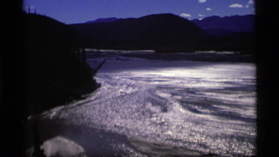 1977 Alaska 8mm frame showing a reflective waterway bordered by dark forested hills and distant mountains.