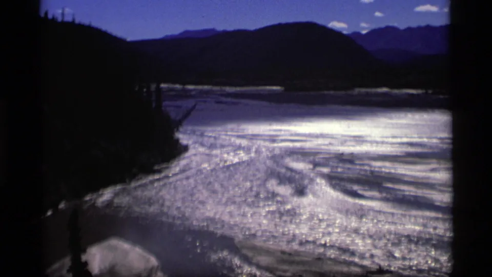 A wide 1977 Alaska home-movie view of bright water, silhouetted shoreline, hills, and distant mountain forms.
