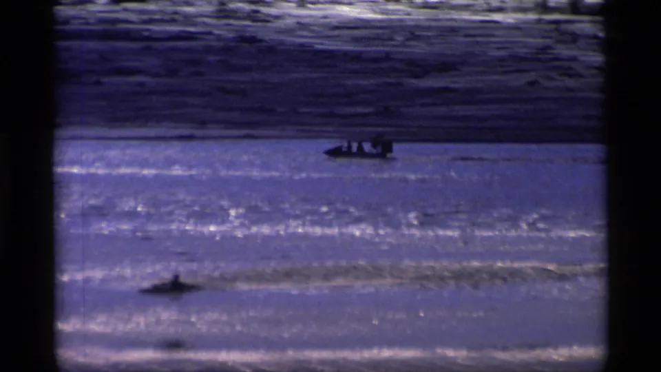 Distant people in a small boat move across a reflective Alaskan waterway in 1977 8mm home-movie footage.