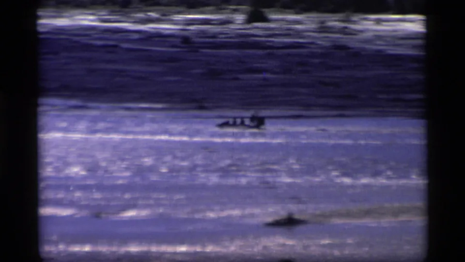Distant boaters and possible birds are visible on a blue Alaskan waterway in this 1977 8mm frame.