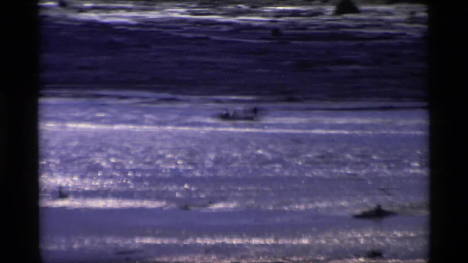 1977 Alaska 8mm frame showing a distant small boat moving across reflective water under dark shoreline bands.