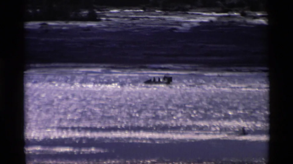 1977 Alaska 8mm frame showing a distant small boat on sparkling water, with a darker shore or hillside behind it.