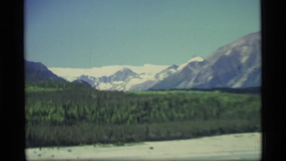 1977 Alaska 8mm frame showing forest, a wide pale riverbank, and snow-covered mountains under a clear sky.