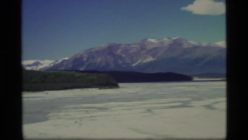 Aerial 8mm frame of an Alaskan frozen lake or river, tree-covered shoreline, and mountain slopes beneath blue sky.