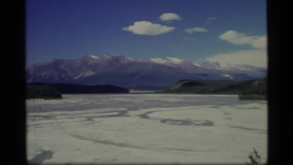 1977 Alaska aerial view of a broad frozen lake with curved surface markings, low forest, and snow-capped mountains beyond.
