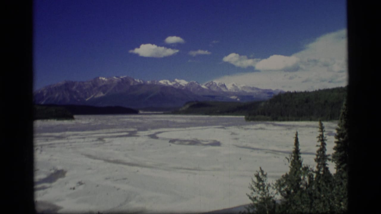 Snow-covered lake or valley below distant mountains with trees at the edge.