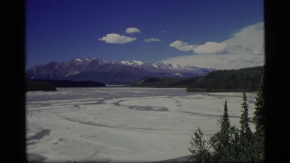 1977 Alaska 8mm frame showing a broad snowy lake or valley, distant mountains, evergreen trees, blue sky, and film borders.