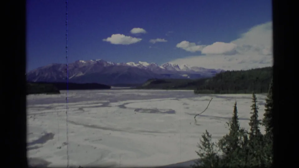 1977 Alaska 8mm view of a snow-covered lake or valley with mountain peaks, forested shore, clouds, and vertical film scratches.