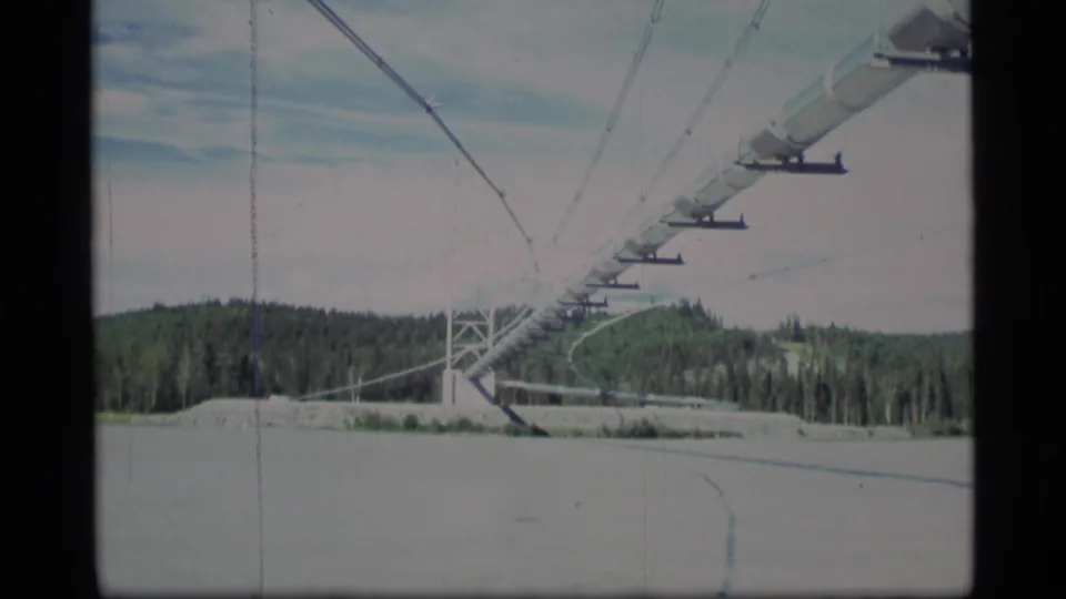 1977 Alaska 8mm frame showing ski-lift cables, support hardware, a snowy ground plane, forested hills, and pale winter sky.