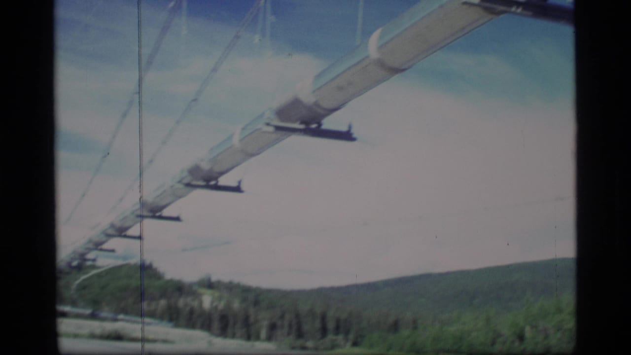 1977 Alaska 8mm frame showing a pale supported pipe or beam stretching across blue sky with forested hills below.