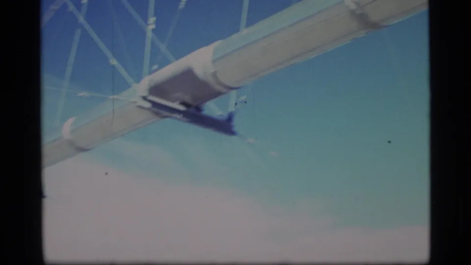 Close 1977 Alaska home-movie view of a pale industrial span and cable supports against a bright blue sky.