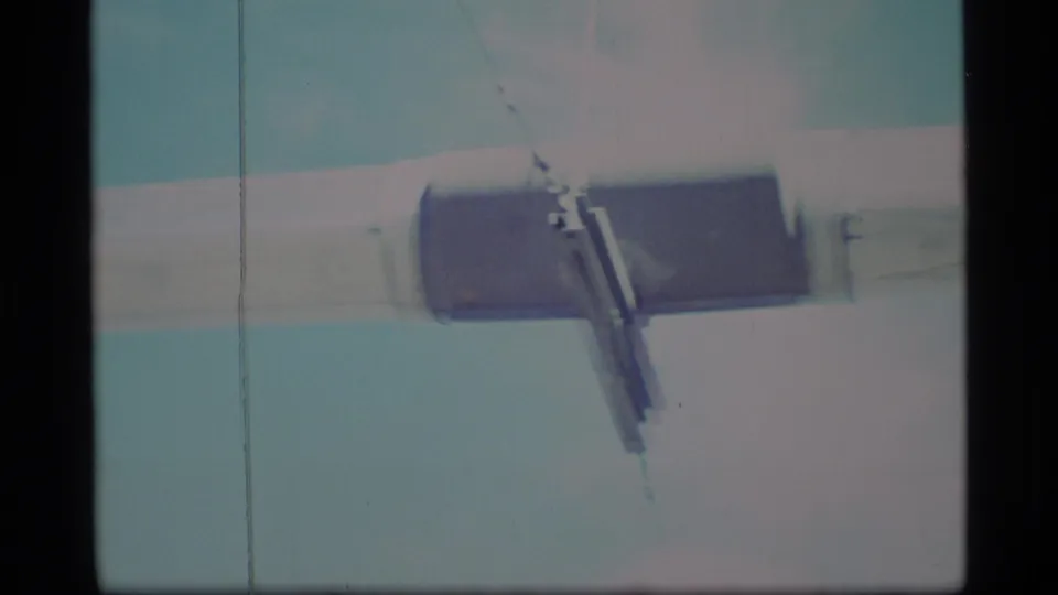 1977 Alaska frame with a close view of a metal bracket attached to a pale beam or pipe under a bright sky.