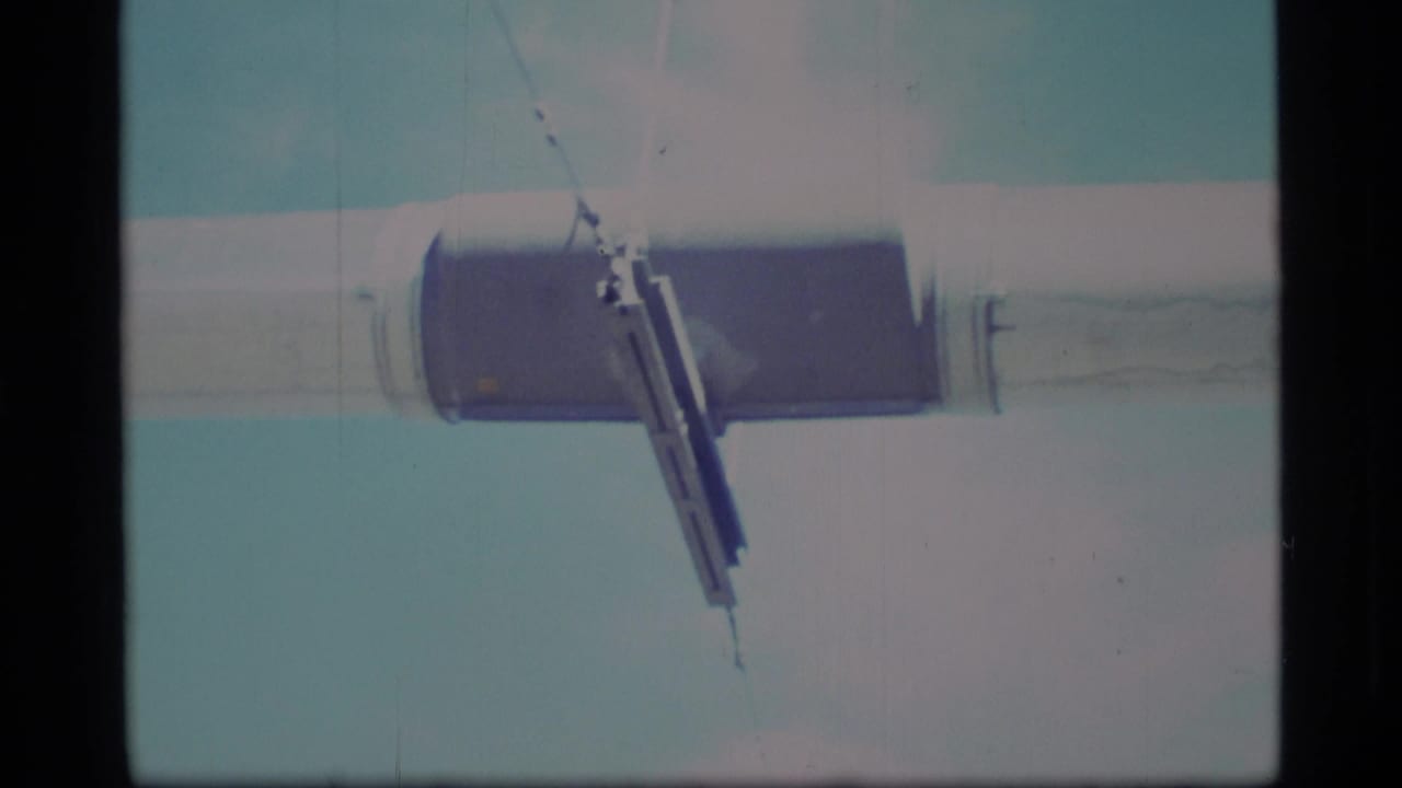1977 Alaska 8mm frame showing a close low-angle view of a silver horizontal beam and dark joint against a pale blue sky.