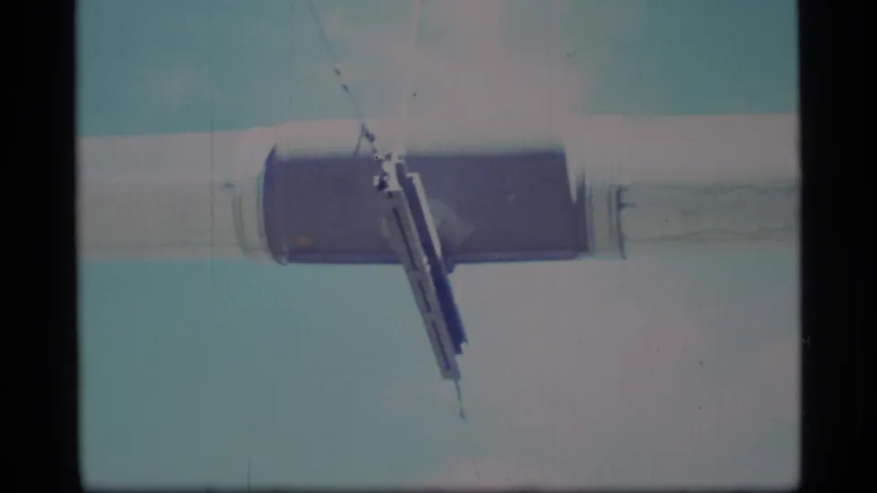 1977 Alaska 8mm frame showing a close low-angle view of a silver horizontal beam and dark joint against a pale blue sky.