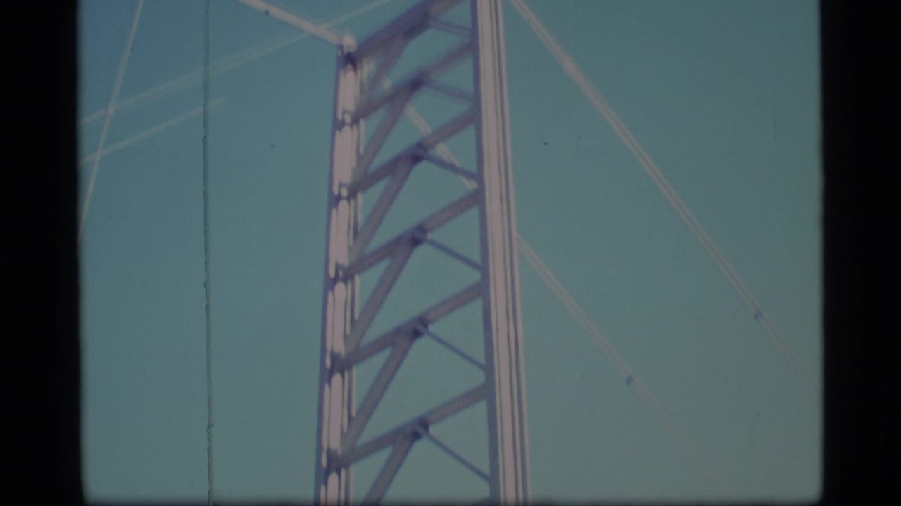 Upper section of a tall metal lattice tower rising against a pale blue sky.