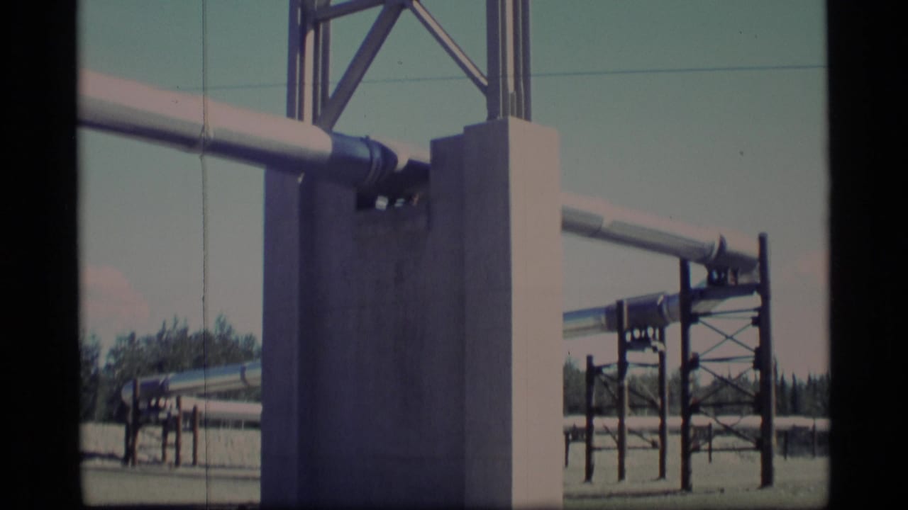 1977 Alaska 8mm frame showing an elevated pipeline supported by concrete piers and metal framing at a remote gravel worksite.