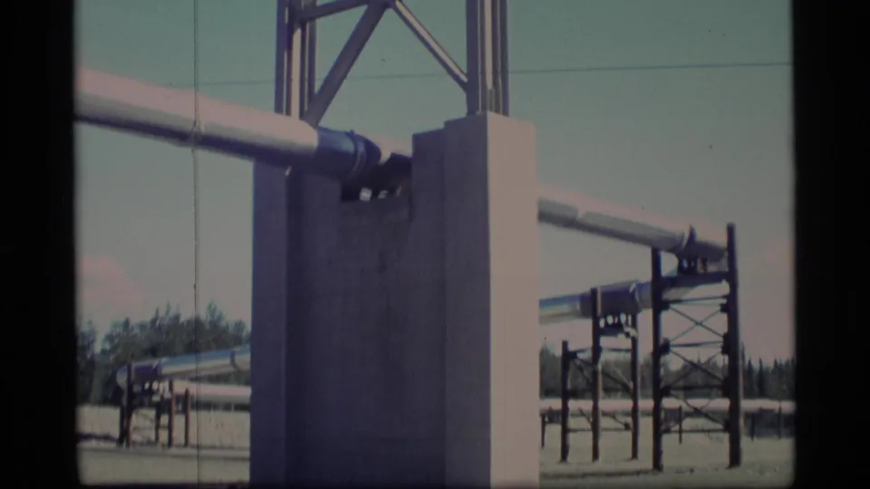 1977 Alaska 8mm frame showing an elevated pipeline supported by concrete piers and metal framing at a remote gravel worksite.
