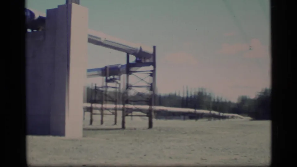 A faded 8mm view of elevated pipeline infrastructure stretching across open ground in Alaska, with forest in the background.