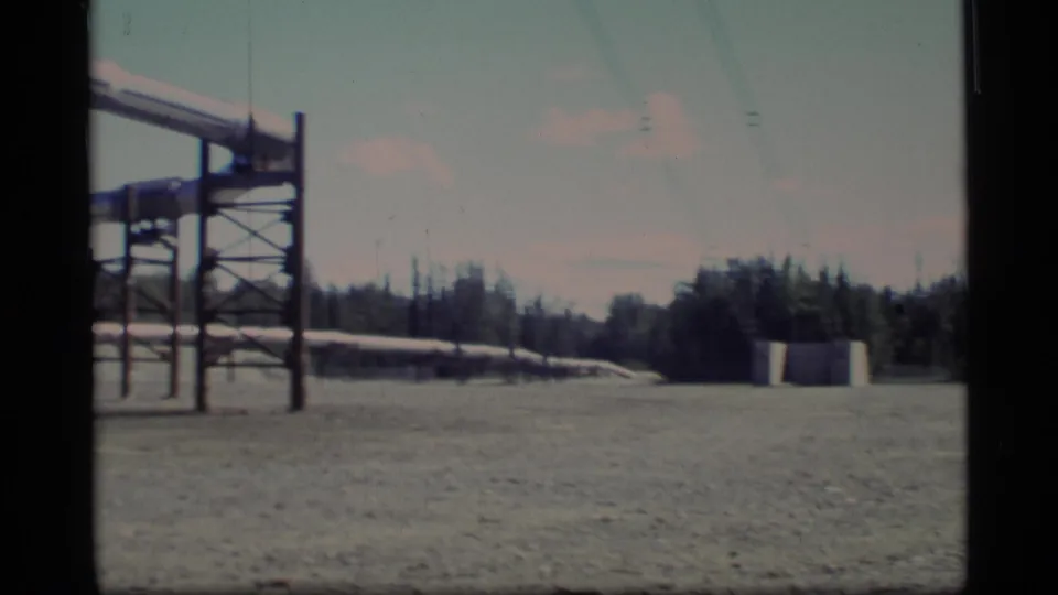 1977 Alaska home-movie frame with pipeline supports at left, a broad gravel worksite, forest, and low concrete forms in the distance.