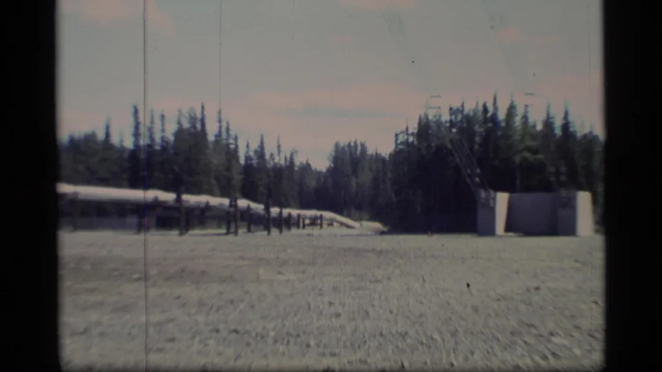 Wide 8mm frame of Alaska pipeline infrastructure crossing a gravel field, with evergreen forest and distant utility towers visible.